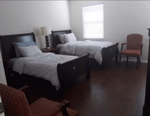 A bedroom with two twin beds dressed in patterned bedding, a nightstand with a lamp, two chairs, and a window.