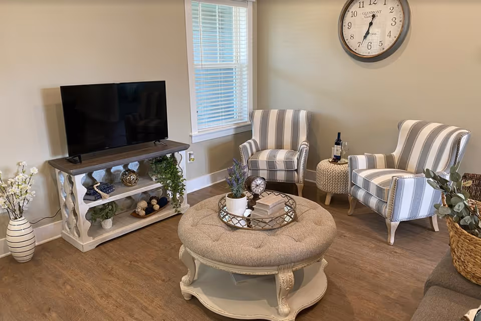 A cozy living room area with two striped armchairs, a round tufted ottoman coffee table with decorative items, a TV on a white wooden stand, a large wall clock, and a window with blinds. The room has wooden flooring and neutral-colored walls.