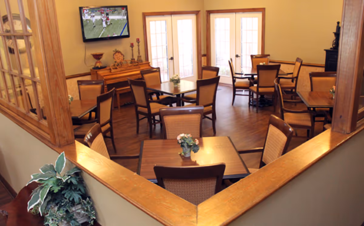 A cozy dining room with several wooden tables and chairs arranged neatly. Each table has a small potted plant centerpiece. There is a piano against the far wall beneath a mounted flat-screen TV showing a football game. The room has wooden flooring, beige walls, and large glass doors letting in natural light.