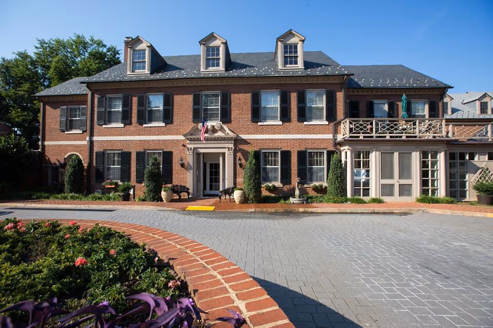Front exterior view of a large, two-story brick building with black shutters and three dormer windows on the roof. The entrance has a small portico with an American flag above it. There are potted plants and greenery around the entrance, and a circular flower bed with red flowers in the foreground. The sky is clear and blue.