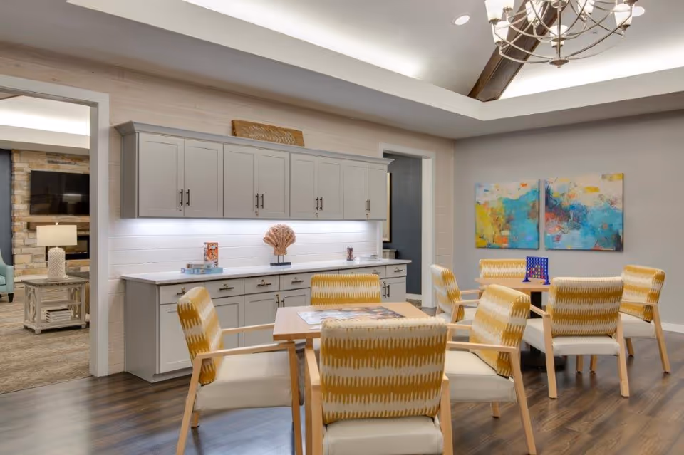 Bright communal dining/activity room with a wooden table surrounded by patterned chairs, built-in cabinets, colorful wall art, and a chandelier.