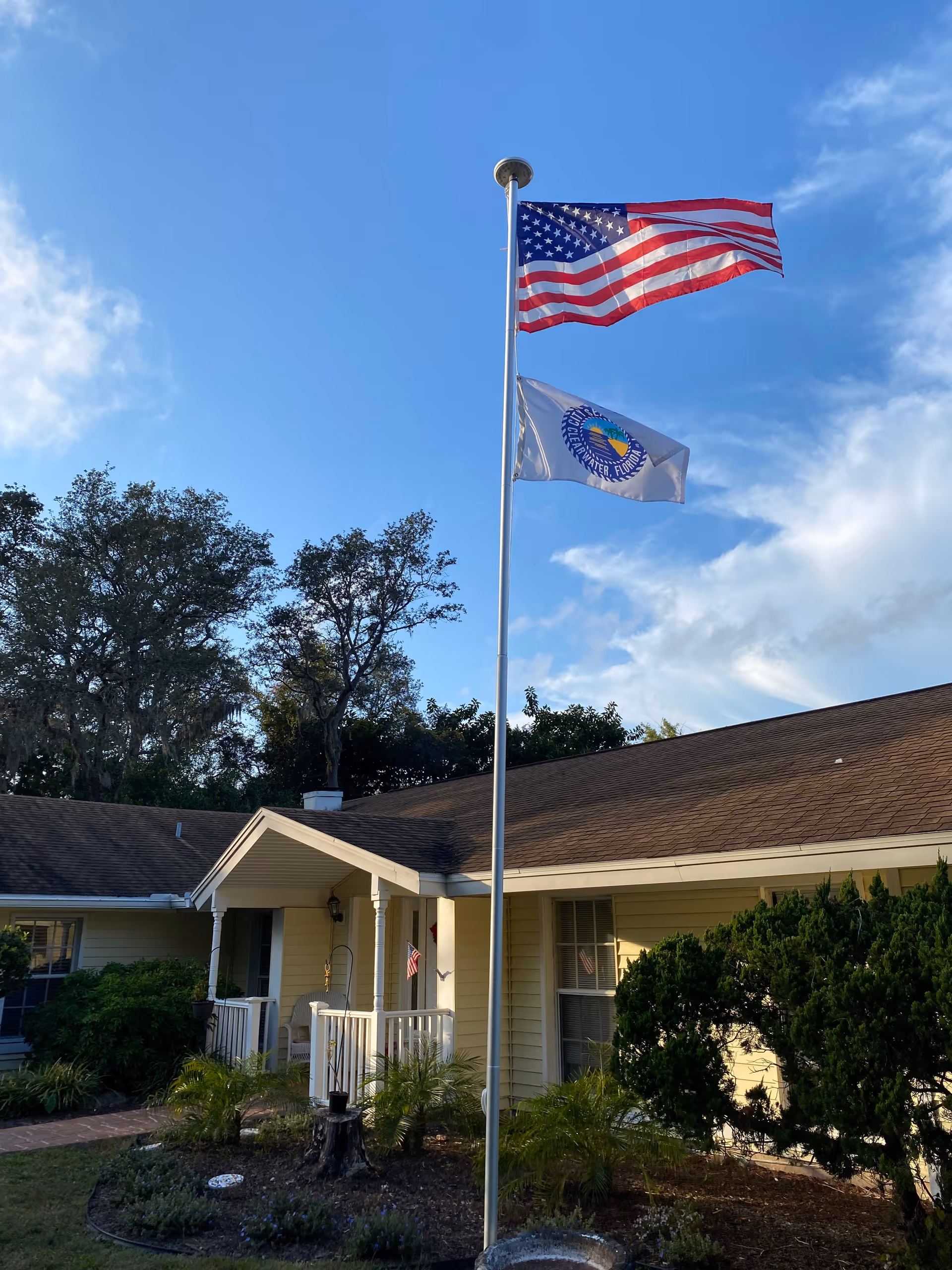 Exterior view of a single-story building with a brown shingled roof and light yellow siding. In front of the building is a flagpole with the American flag and another flag below it. There are trees and shrubs around the building, and the sky is partly cloudy with blue patches.