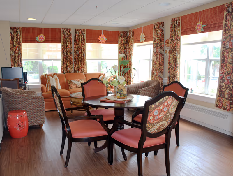 A cozy living room area with a round wooden table surrounded by four upholstered chairs with patterned backs and red seats. Behind the table, there is an orange sofa with patterned cushions and two wicker armchairs. The room has large windows with patterned curtains and red valances, letting in natural light. A decorative vase with flowers is placed on the table. The floor is wooden, and the ceiling has recessed lighting.