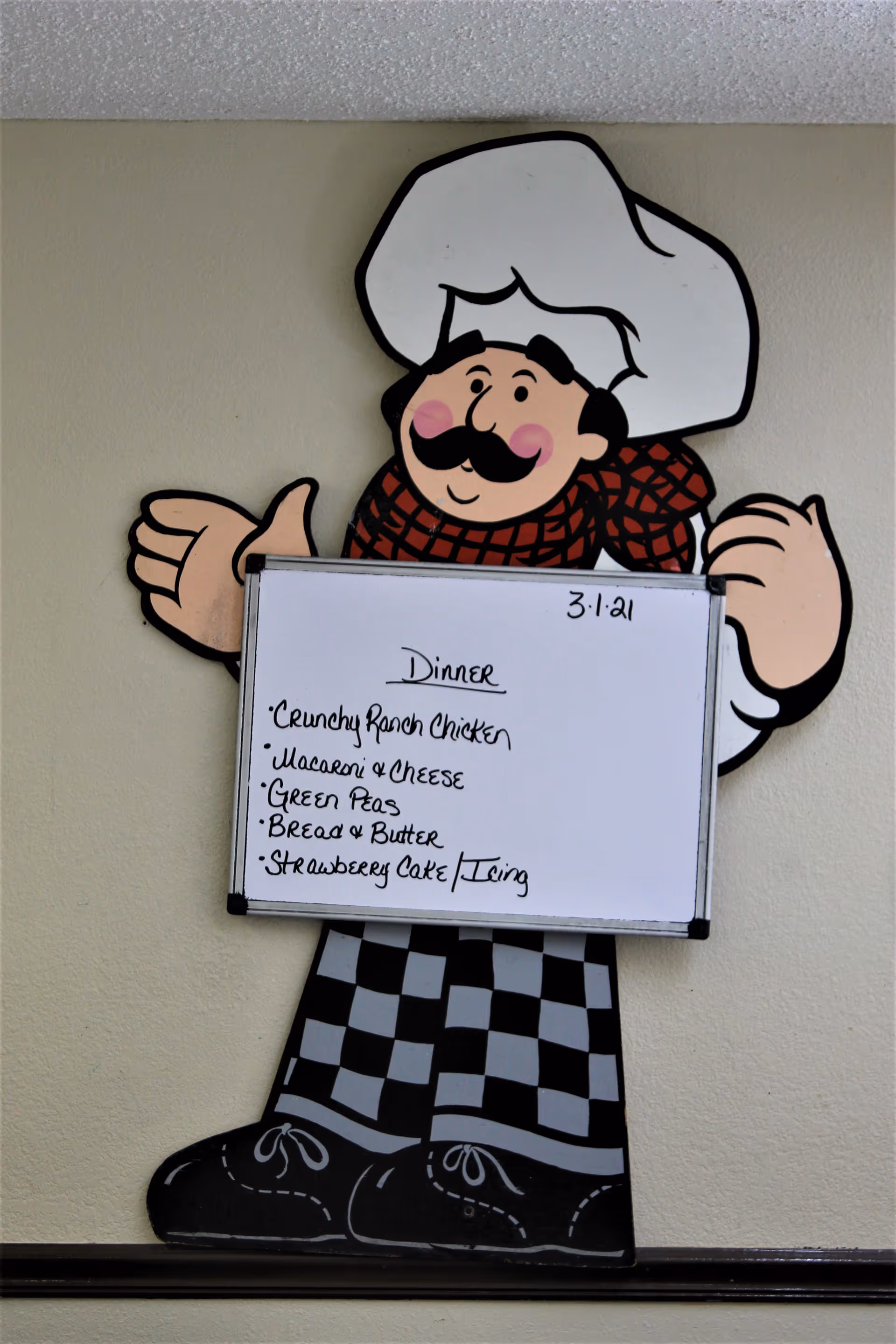 A cartoon chef figure mounted on a wall holding a whiteboard with the dinner menu written on it. The menu includes crunchy ranch chicken, macaroni and cheese, green peas, bread and butter, and strawberry cake with icing. The date 3-1-21 is written at the top right of the whiteboard.