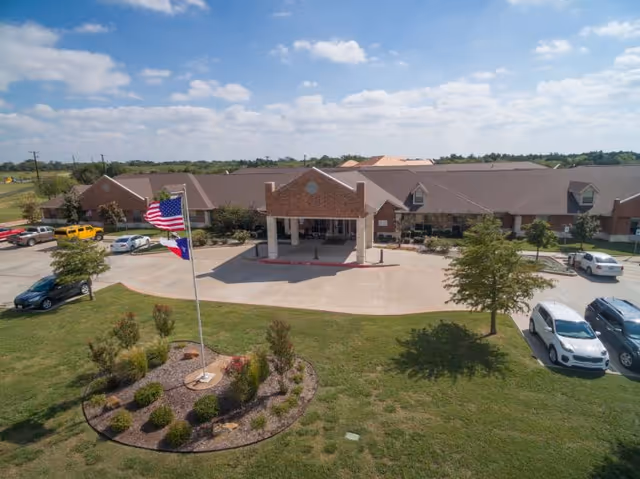 Front exterior view of Legend Oaks Healthcare and Rehabilitation - Ennis building with a covered entrance, a circular driveway, an American flag and Texas state flag on a flagpole in a landscaped area, several parked cars, and a partly cloudy sky.