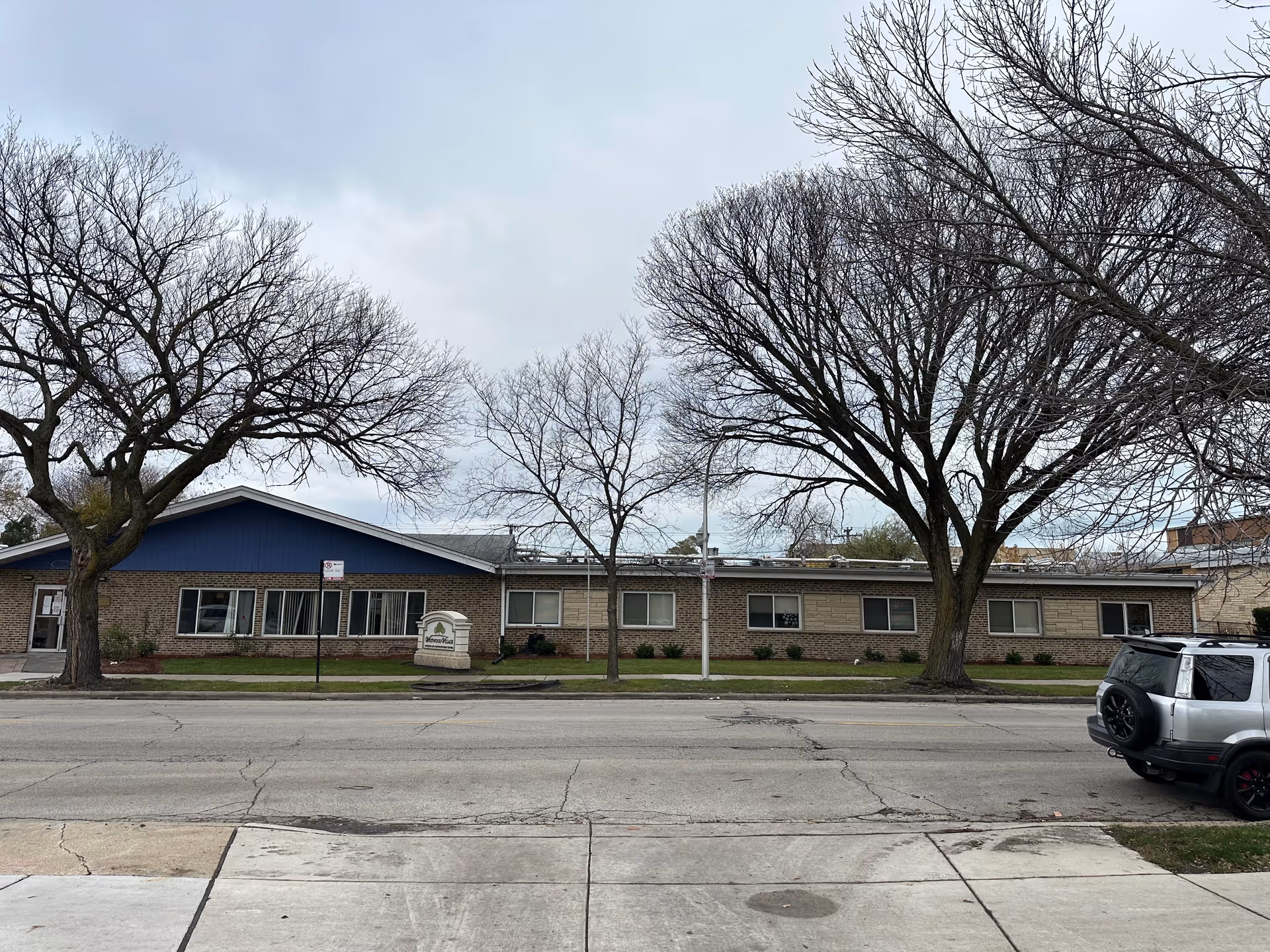 Front view of a single-story brick nursing facility with leafless trees, a sidewalk, street, and a small entrance sign.