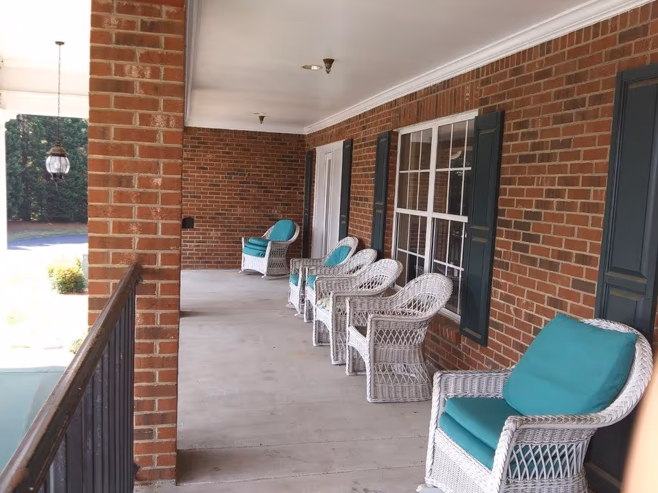 A covered outdoor porch area with a brick wall and several white wicker chairs with teal cushions lined up along the wall. There is a black railing on the left side and a hanging lantern light fixture near the entrance.