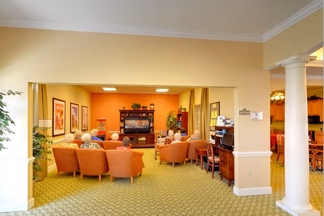 A group of elderly people seated in a cozy common area with orange chairs facing a large television on a wooden entertainment center. The room has warm lighting, framed artwork on the walls, green carpet, and plants. There is a desk with a computer on the right side and a kitchen area visible in the background.
