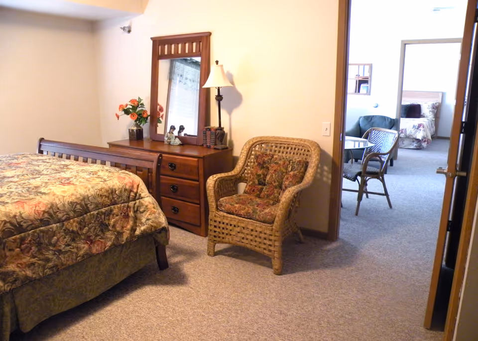 A cozy bedroom with a floral patterned bedspread on a wooden bed frame. Next to the bed is a wooden dresser with a mirror, a table lamp, and decorative items including a vase with flowers. A wicker armchair with floral cushions is placed near the dresser. Through an open doorway, another room is visible with a similar bed, a green armchair, and a wicker chair around a small table.