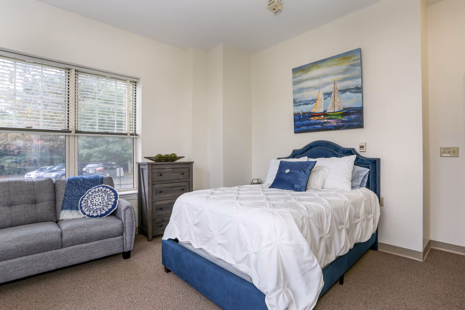Sunlit bedroom featuring a blue upholstered bed with white bedding, a gray sofa by a window, a dresser, and a sailboat painting on the wall.