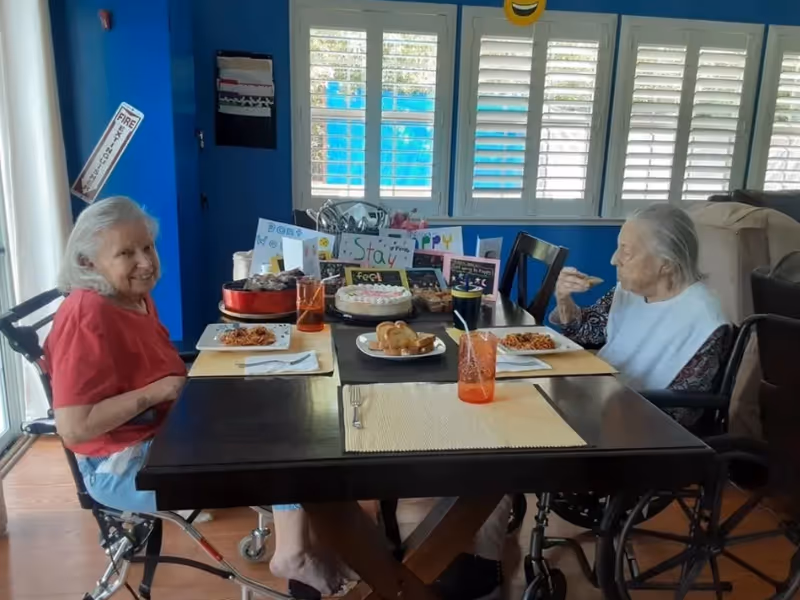 Two elderly women sitting in wheelchairs at a dining table with plates of food and drinks. The table has placemats, a cake, muffins, and colorful handmade signs in the background. The room has blue walls and large windows with white shutters.