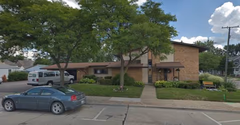 Front exterior of a single-story brick building with trees, parked cars, and a sidewalk.