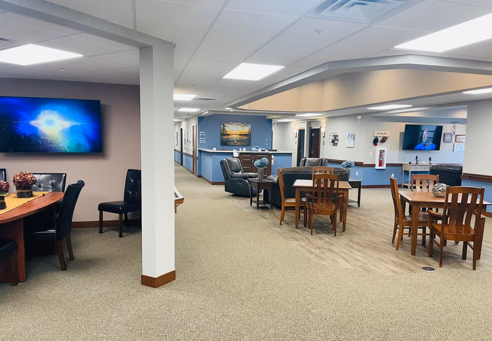 Interior view of a senior living facility common area with multiple seating arrangements including wooden tables with chairs, leather armchairs, and a large TV mounted on the wall. The space has a mix of carpet and wood flooring, soft lighting, and decorative elements like flowers and wall art.