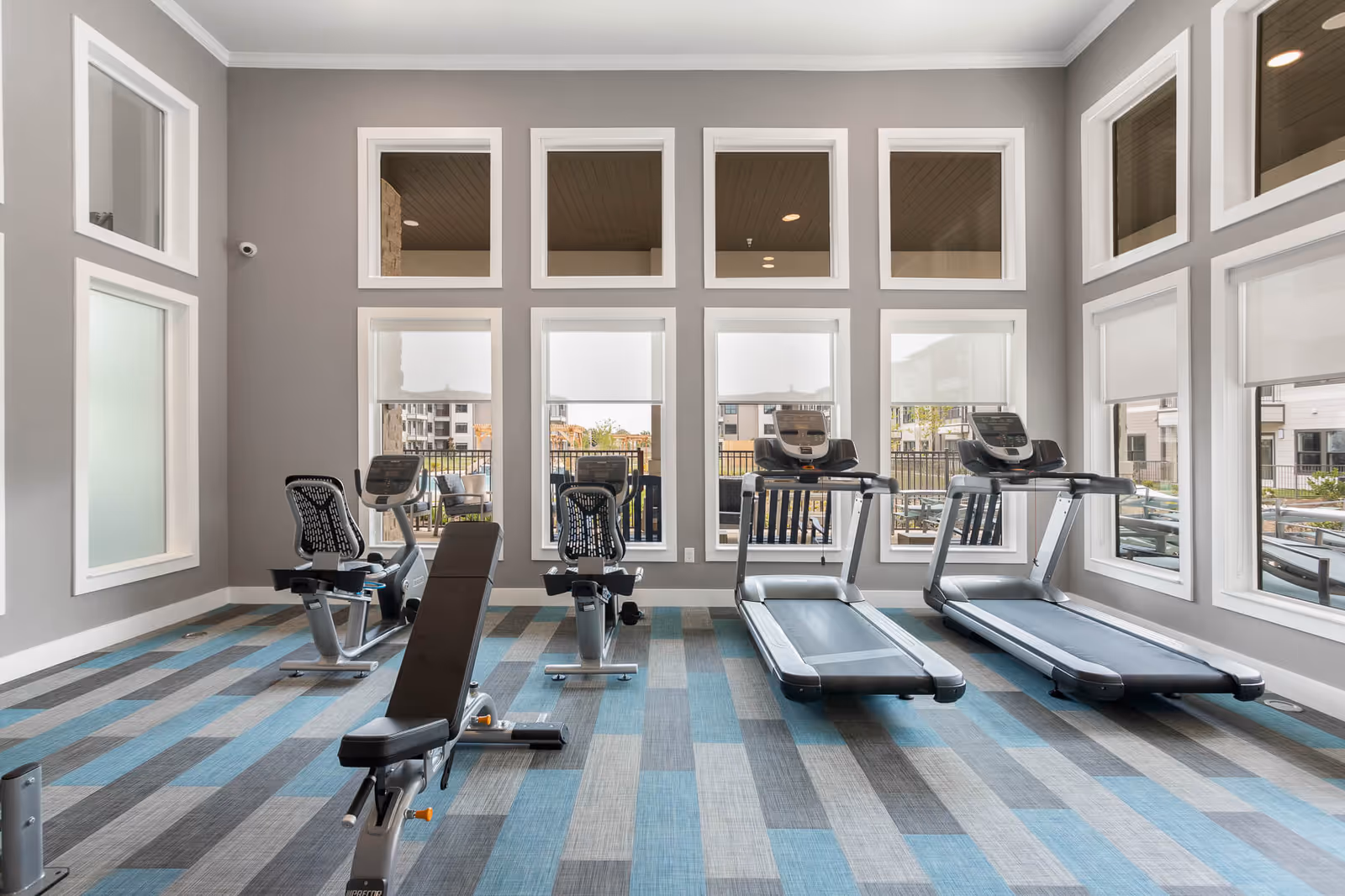 A bright fitness room with large windows and gray walls, featuring two treadmills, two stationary bikes, and a weight bench on a patterned blue and gray carpeted floor.