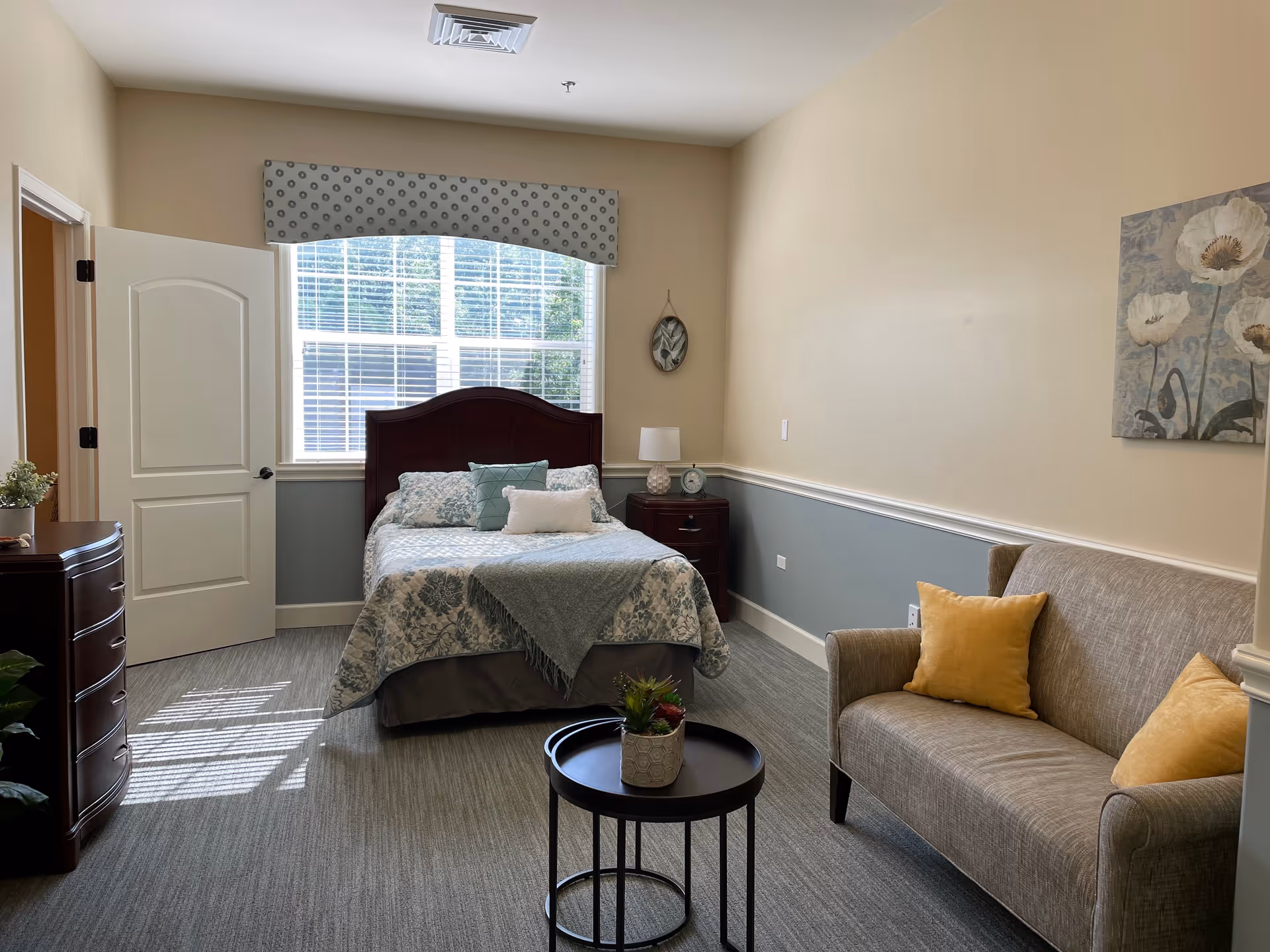A cozy bedroom in a senior living facility featuring a bed with floral bedding and pillows, a wooden headboard, a nightstand with a lamp and clock, a small round table with a potted plant, and a beige loveseat with two yellow pillows. The room has a large window with blinds and a patterned valance, light-colored walls with a chair rail, and a floral painting on the wall.