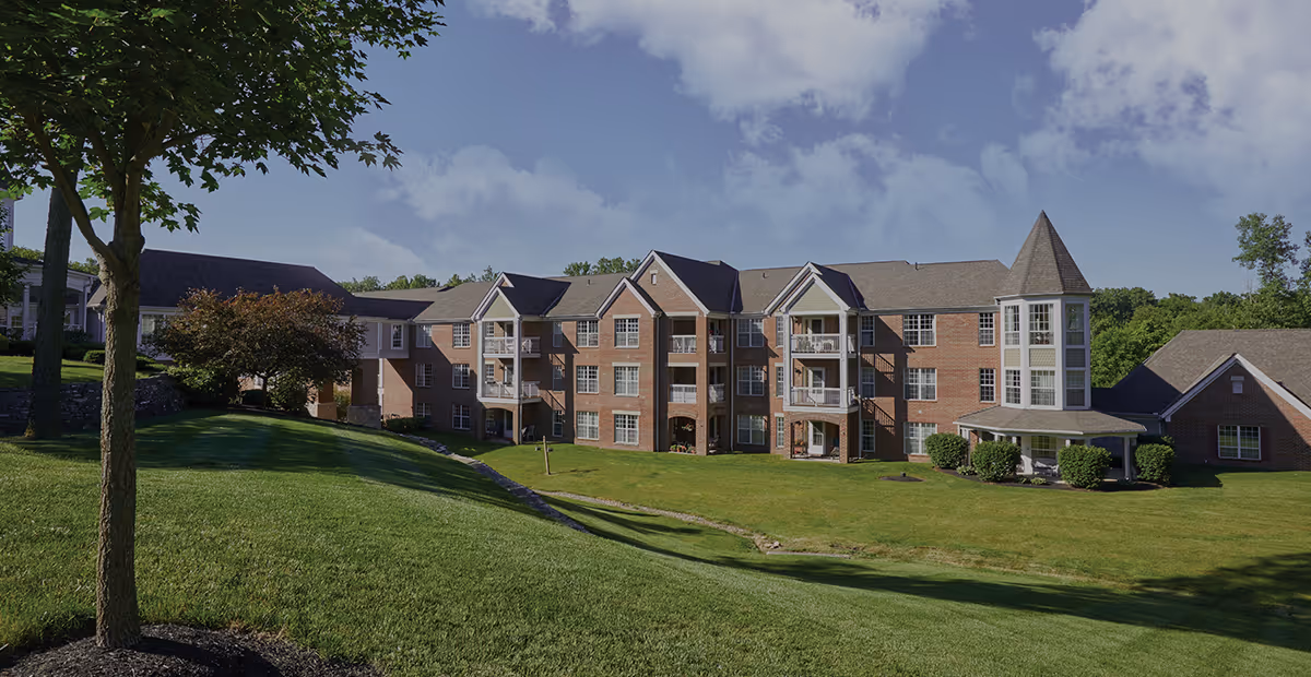 A large, multi-story brick building with balconies and a turret-like structure on the right side, surrounded by a well-maintained lawn and trees under a partly cloudy sky.