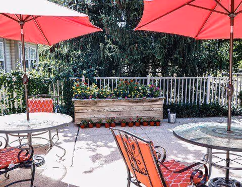 Sunny outdoor patio with red umbrellas, metal tables and chairs, and a wooden planter full of flowers by a railing.