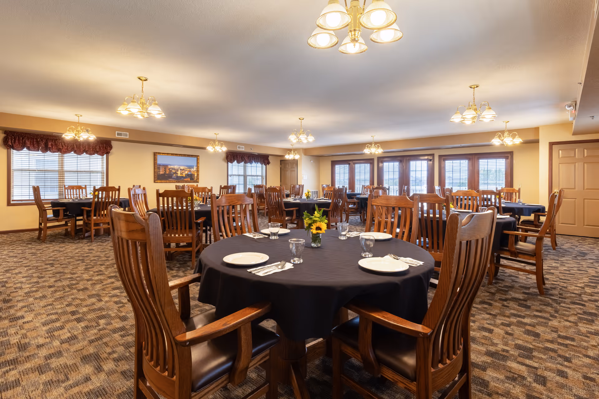 A spacious dining room with multiple round and rectangular wooden tables covered with black tablecloths. Each table is set with white plates, glasses, and silverware. The room has carpeted flooring, beige walls, several windows with blinds and valances, and multiple ceiling light fixtures providing warm lighting.