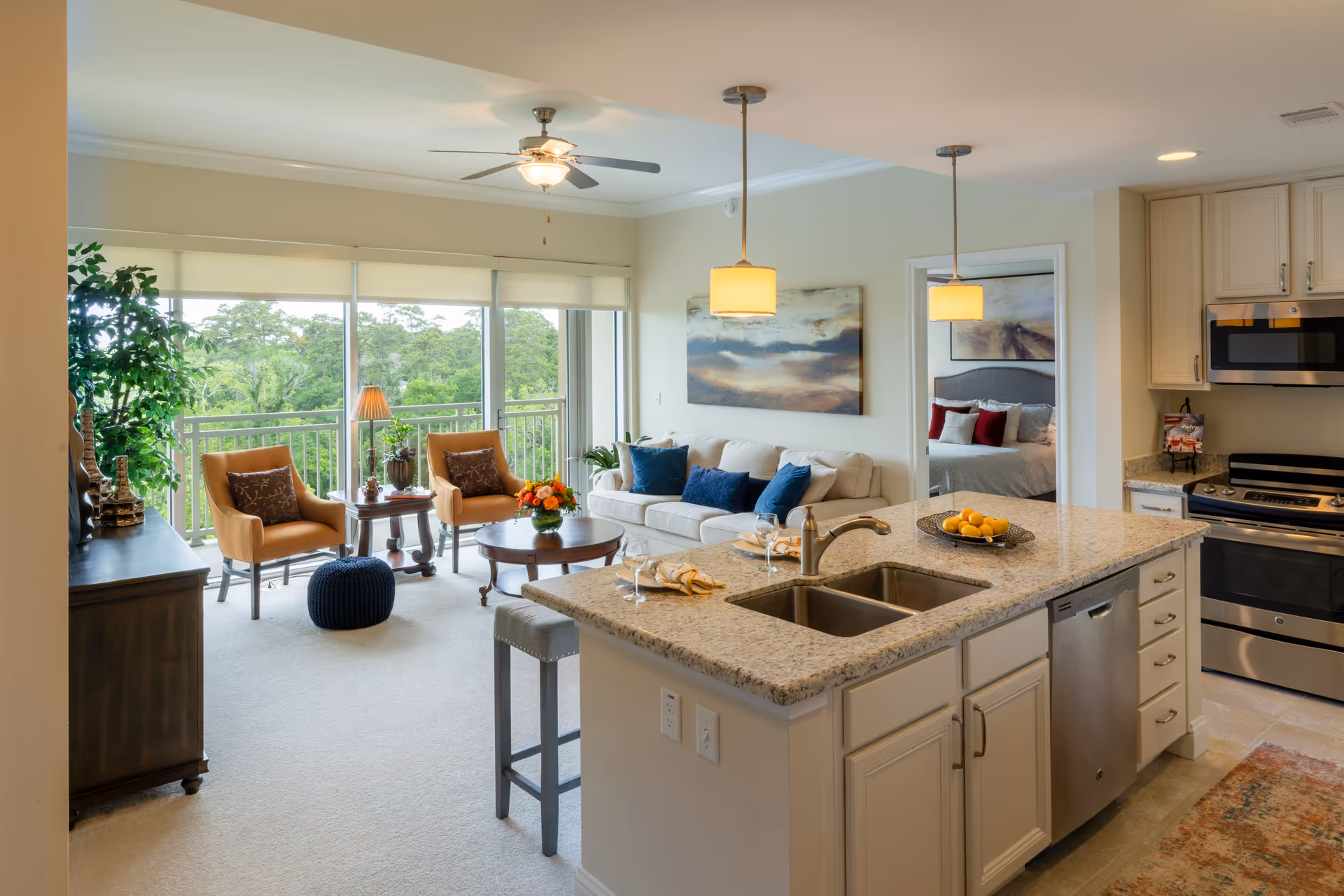 Bright and spacious open-concept living area in a senior living facility featuring a kitchen with granite countertops, stainless steel appliances, and a double sink. The living room has a white sofa with blue cushions, two tan armchairs, a round wooden coffee table with a flower arrangement, and large windows with a view of greenery outside. A ceiling fan and pendant lights illuminate the space. A bedroom with a neatly made bed and artwork is visible through an open doorway.
