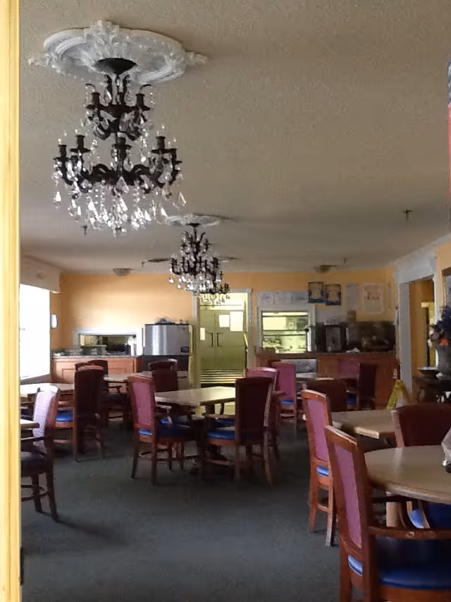 Interior view of a dining room with multiple round tables and chairs arranged around them. The room has yellow walls and green carpet flooring. Two ornate chandeliers hang from the ceiling. In the background, there is a kitchen area with appliances and cabinets.