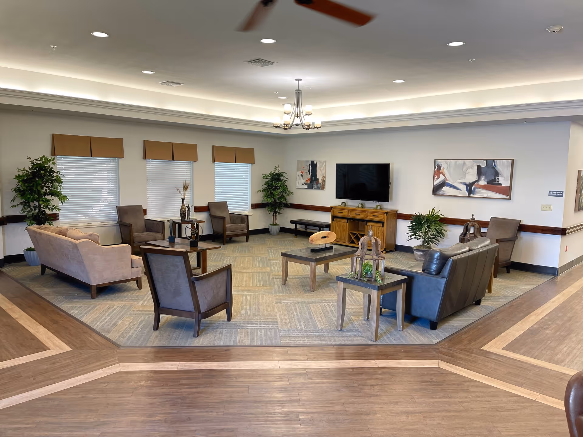 A spacious and well-lit living room area with multiple seating options including beige and dark brown sofas, and several armchairs arranged around two coffee tables. The room features large windows with blinds, potted plants, a flat-screen TV mounted on the wall above a wooden console, and modern ceiling lighting with a fan.
