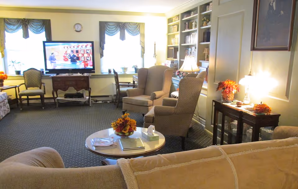 A cozy living room with beige upholstered armchairs and a sofa arranged around a round coffee table with a flower arrangement and books. A TV is on a wooden stand against the wall with two windows behind it, and there are additional chairs and a dining table near the windows. The room is warmly lit with table lamps and decorated with flowers and framed artwork.