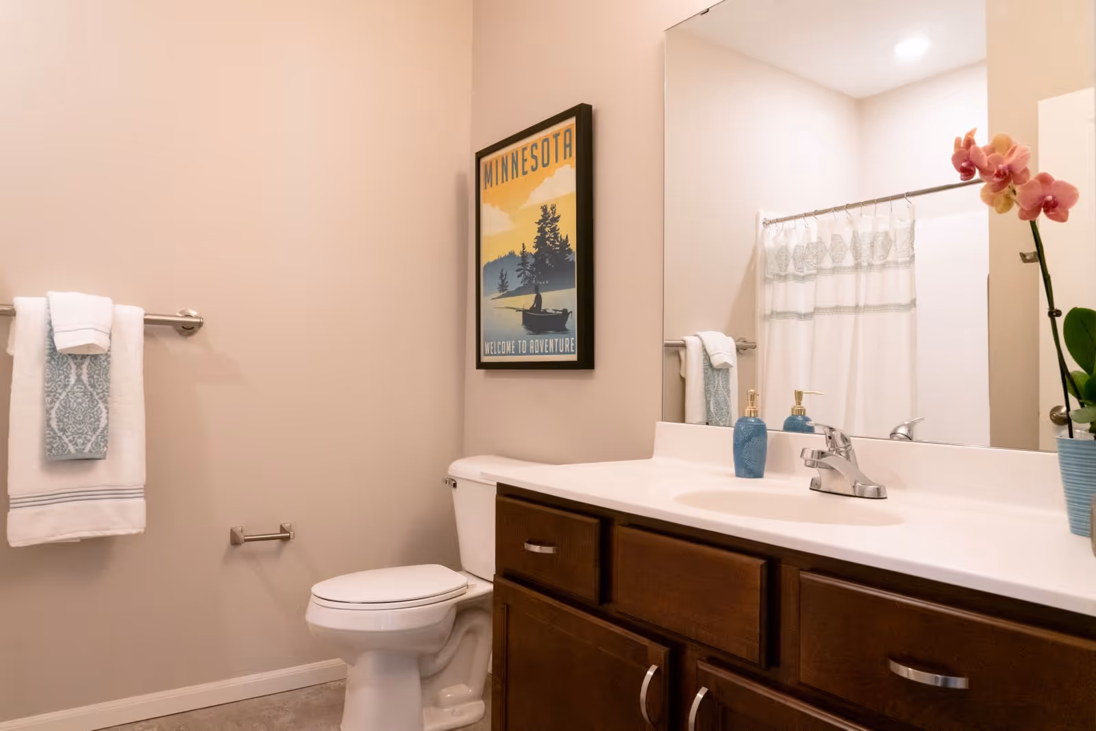 A clean bathroom featuring a white toilet, a wooden vanity with a white countertop and sink, a large mirror above the sink, a towel rack with folded towels, a shower curtain with a decorative pattern, a framed Minnesota-themed poster on the wall, and a potted orchid plant on the countertop.