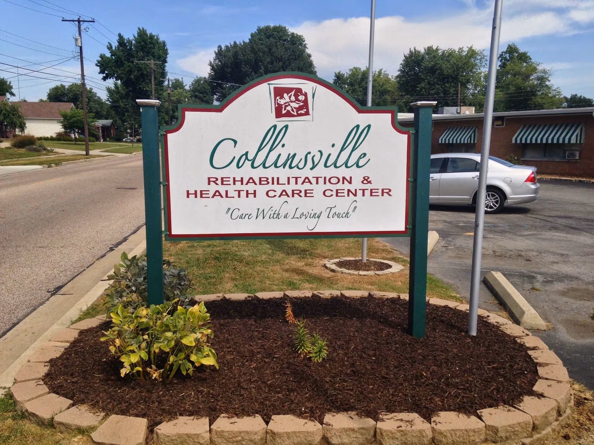Outdoor view of a sign for Collinsville Rehabilitation & Health Care Center with the slogan 'Care With a Loving Touch'. The sign is mounted on green posts and surrounded by a small landscaped area with mulch and plants. In the background, there is a parking lot with a silver car and a building with green and white striped awnings.
