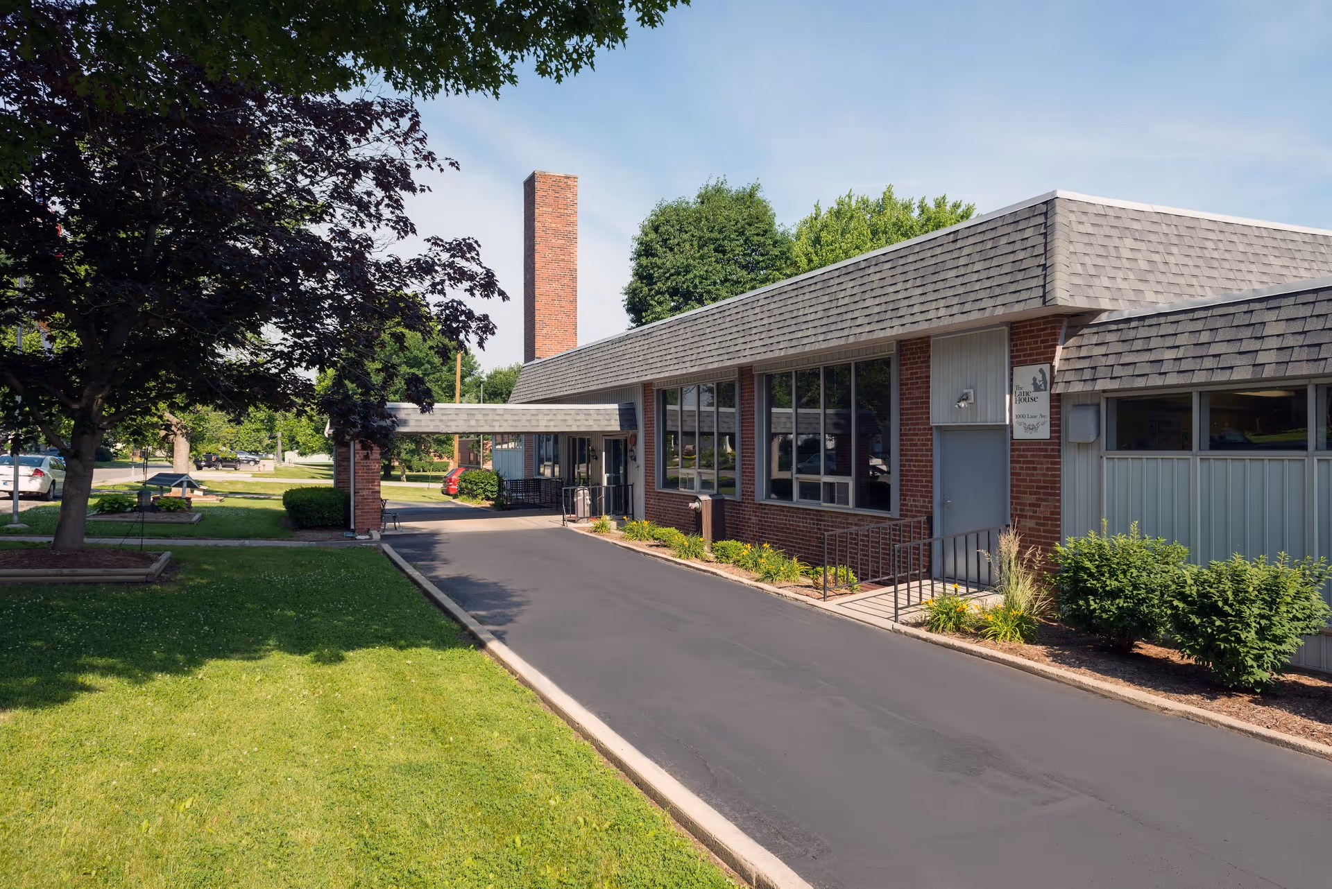 Exterior view of The Lane House facility showing a single-story brick building with large windows, a covered entrance, a paved driveway, and well-maintained green lawn and trees under a clear sky.