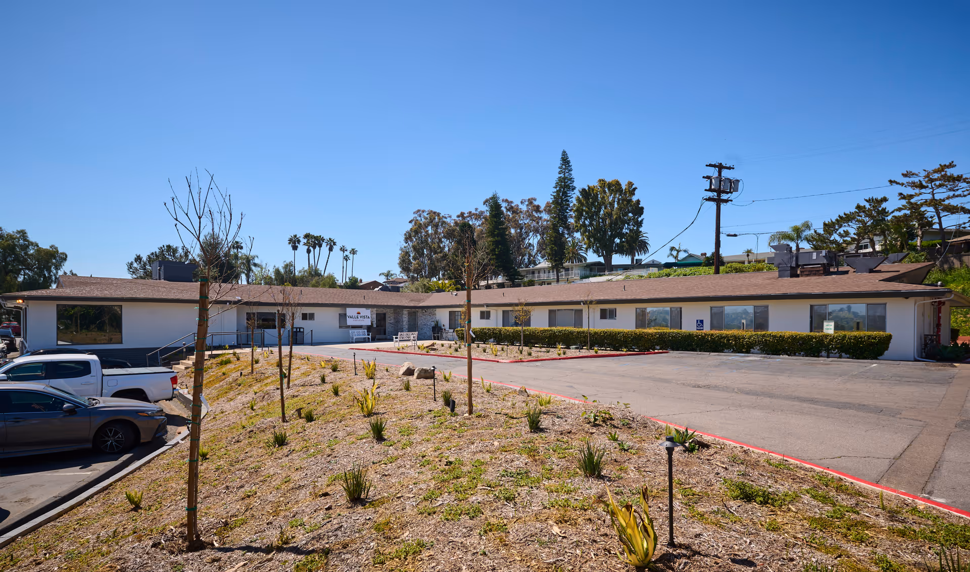 Exterior view of Valle Vista Post Acute facility showing a single-story building with a brown roof, several windows, and a parking area with a few parked cars. The foreground has a landscaped area with small plants and young trees under a clear blue sky.
