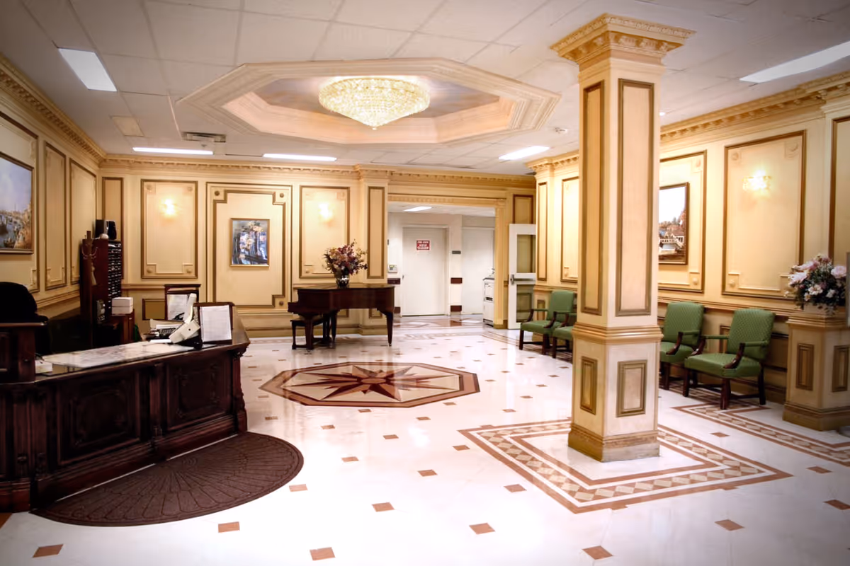 Spacious elegant reception lobby with a wooden front desk, green chairs, decorative pillars, and a central chandelier.