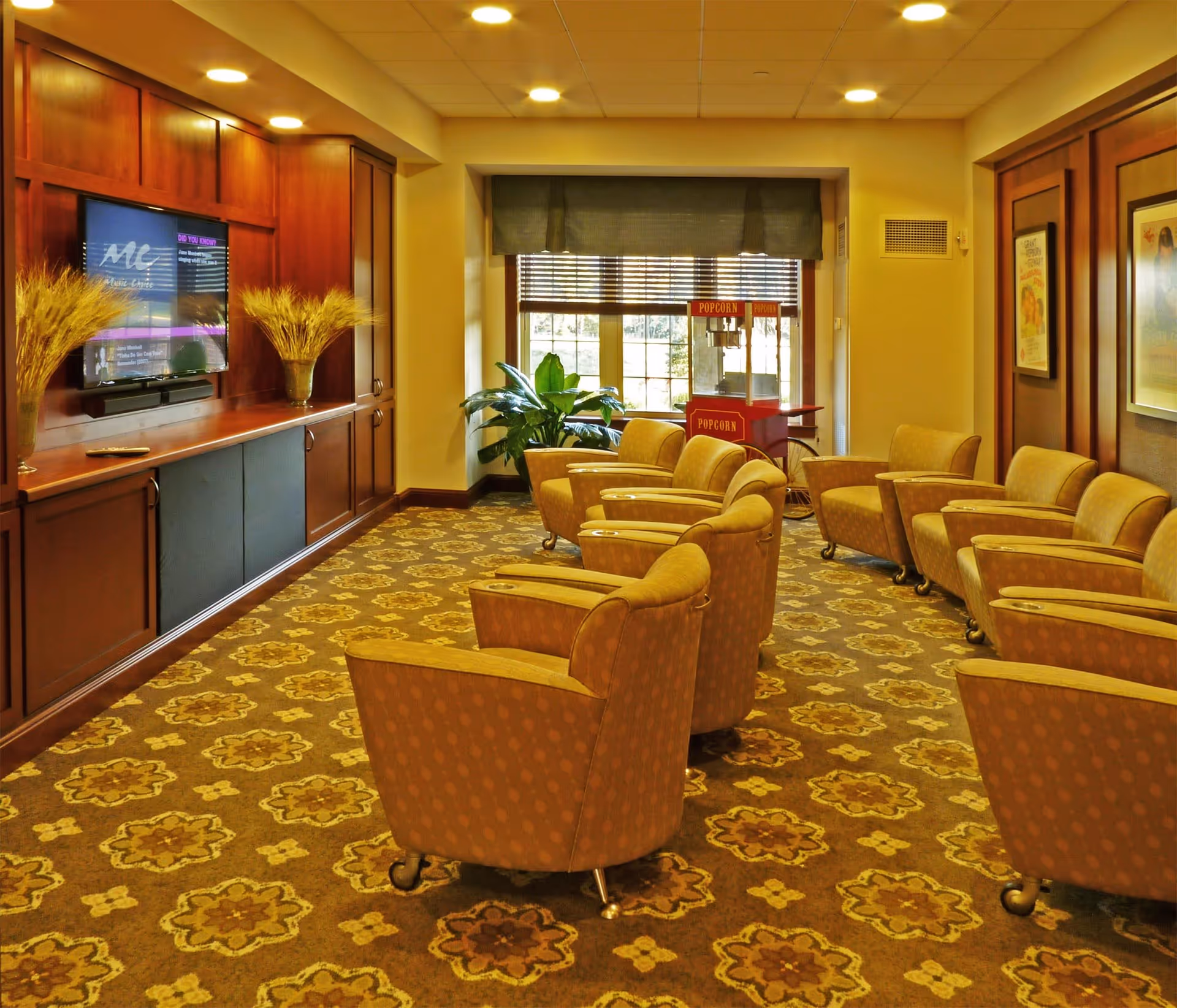 A cozy assisted living common area with patterned carpet, several upholstered armchairs arranged in rows facing a wall-mounted TV, wooden cabinetry, a popcorn machine near a window with blinds, and framed artwork on the walls.