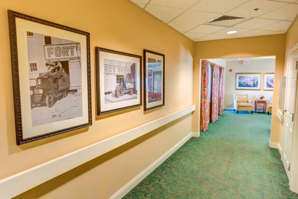 A hallway in a senior living facility with beige walls and green carpet. Three framed black and white vintage photographs hang on the left wall. At the end of the hallway, there is a small seating area with two beige armchairs, a wooden side table with a flower vase, and two framed pictures on the wall. Colorful curtains partially cover a doorway on the right side near the seating area.