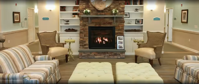 A cozy living room area with a stone fireplace in the center, flanked by built-in white shelves decorated with flowers and small sculptures. There are two beige armchairs with wooden legs on either side of the fireplace, a striped sofa on the left, and two tufted ottomans in front. The walls are painted light green with white wainscoting, and there are framed pictures and wall sconces providing soft lighting.
