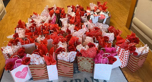 Numerous Valentine's-themed gift bags with heart patterns and red and pink tissue paper arranged on a wood floor in an interior hallway.
