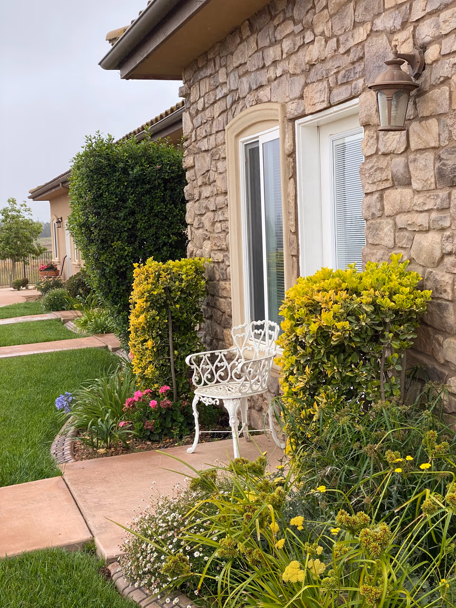 Outdoor view of a stone building wall with a white decorative metal chair placed on a concrete pathway. The area is surrounded by green grass, various bushes, and flowering plants. A wall-mounted lantern light is visible next to a window and door with blinds.