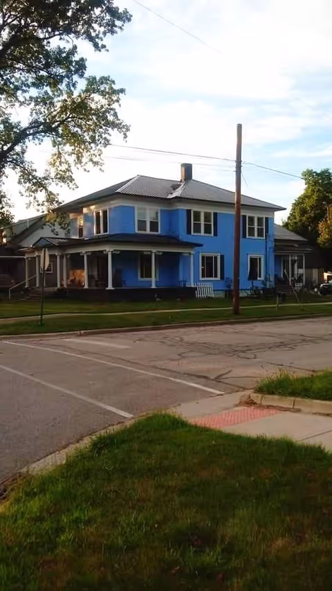 A two-story blue house with white trim and a covered porch, situated on a corner lot with green grass and trees nearby. The house has multiple windows and a metal roof. There is a street and sidewalk in the foreground.