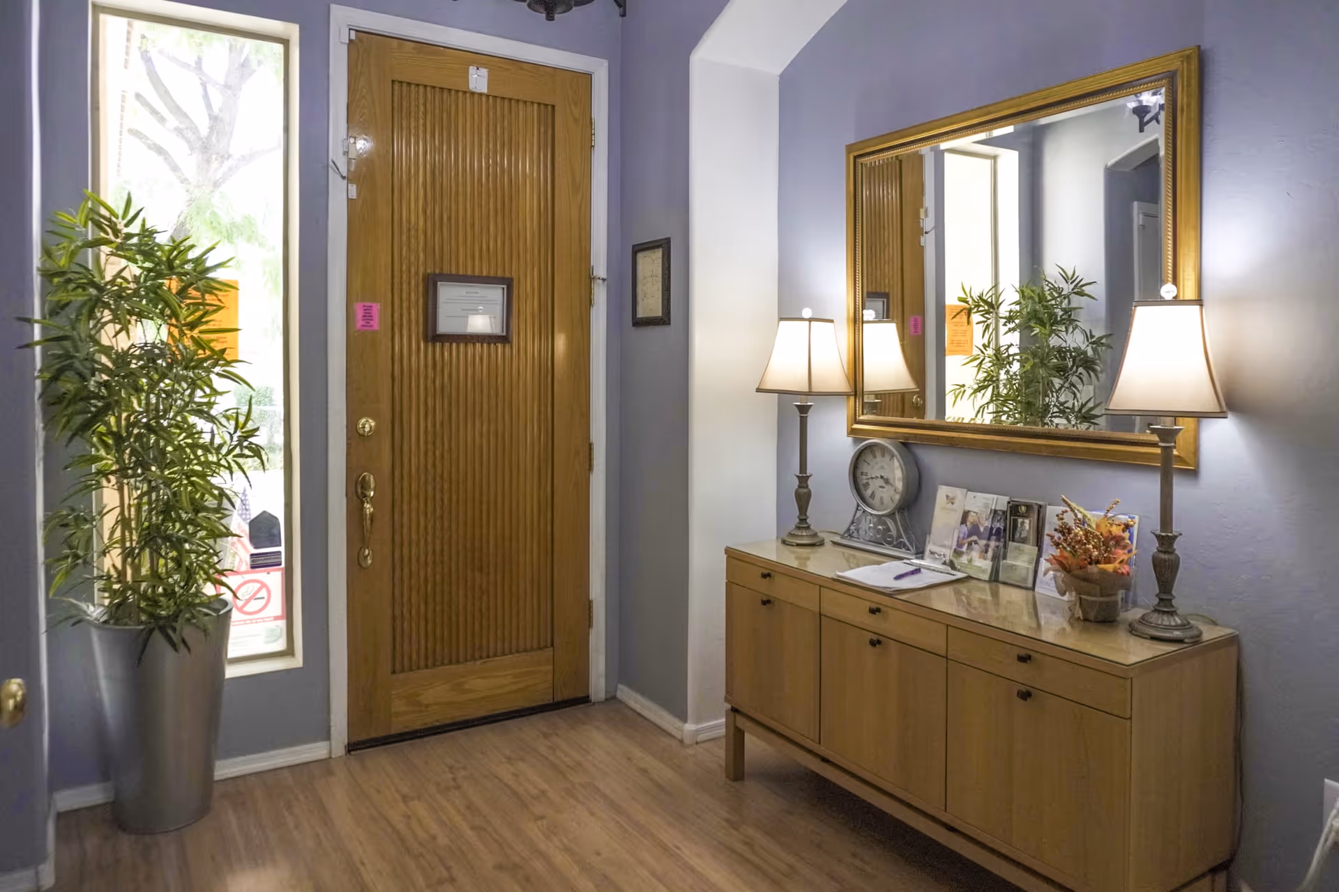 Entryway interior with a wooden front door, tall side window, potted plant, and a console table topped by lamps, a mirror, and decorative items.