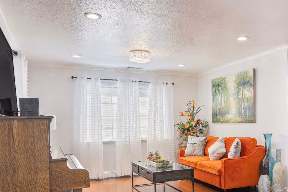 A bright living room with a large window covered by sheer white curtains. There is an orange sofa with three cushions, a glass coffee table with a small plant, a wooden piano on the left side, and a painting of trees on the wall above the sofa. Decorative vases and a floral arrangement are also visible.