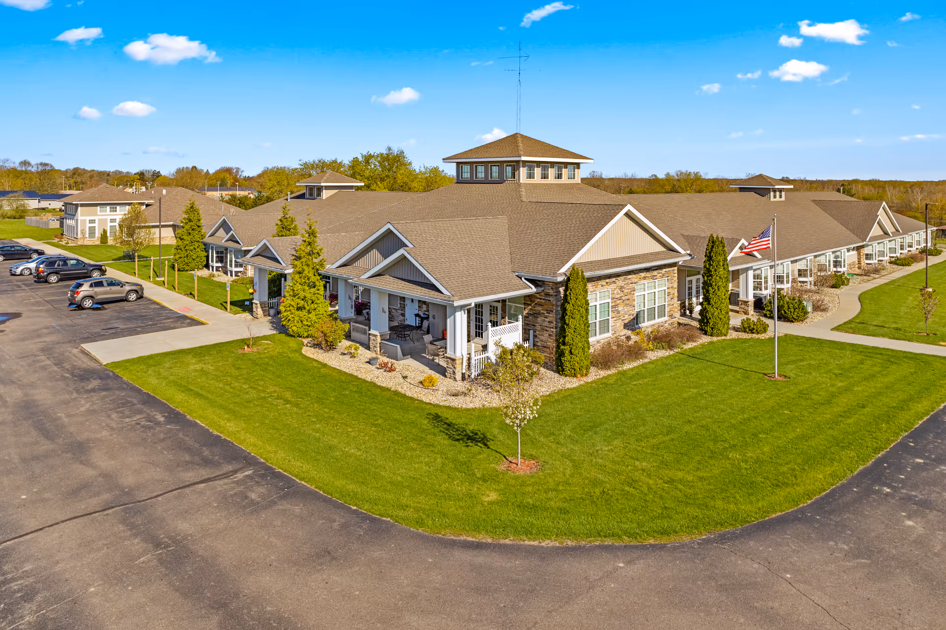 Aerial view of Woodland Terrace of Paw Paw Lake senior living facility showing a large, single-story building with multiple peaked roofs, surrounded by well-maintained green lawns, small trees, and a parking lot with several cars. An American flag is displayed on a flagpole near the building entrance under a clear blue sky.