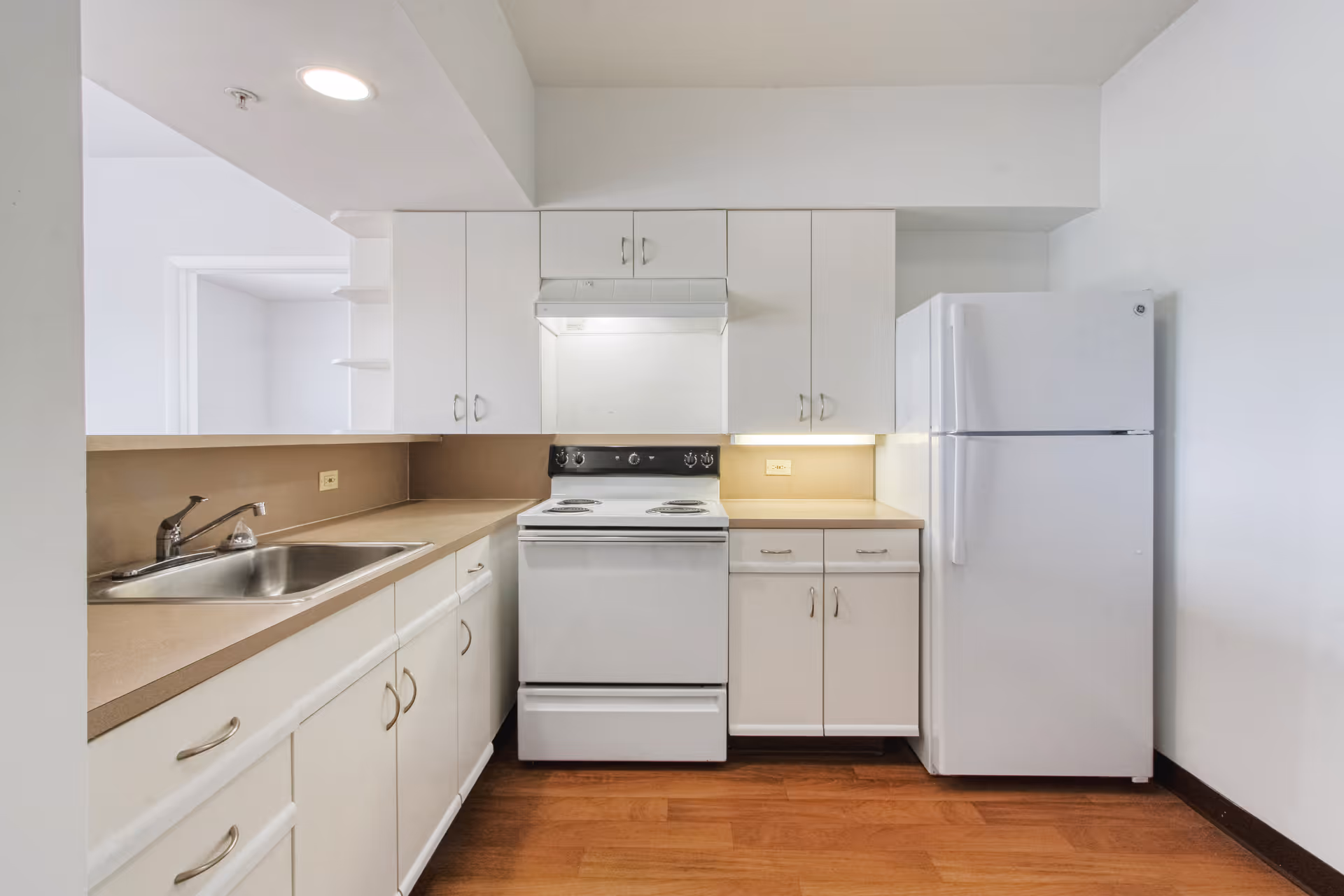 A clean and simple kitchen with white cabinets, a white refrigerator, a white electric stove with an overhead vent, a stainless steel sink, beige countertops, and wood flooring.