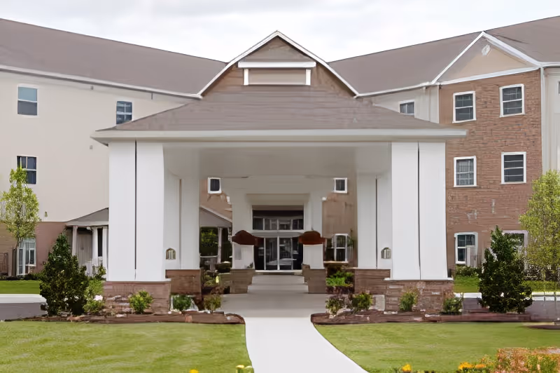 Front exterior view of Sanctuary Grande facility showing a covered entrance with white columns and a brown roof, surrounded by green lawn and small shrubs.