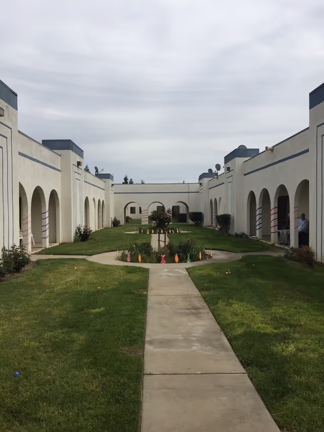 A courtyard area at St. Thomas Retirement Center with a concrete walkway leading through a grassy lawn. The courtyard is surrounded by white buildings with arched walkways and blue trim. There are small bushes and decorative elements along the walkway and in the center garden area. Two people are sitting under the arches on the right side.