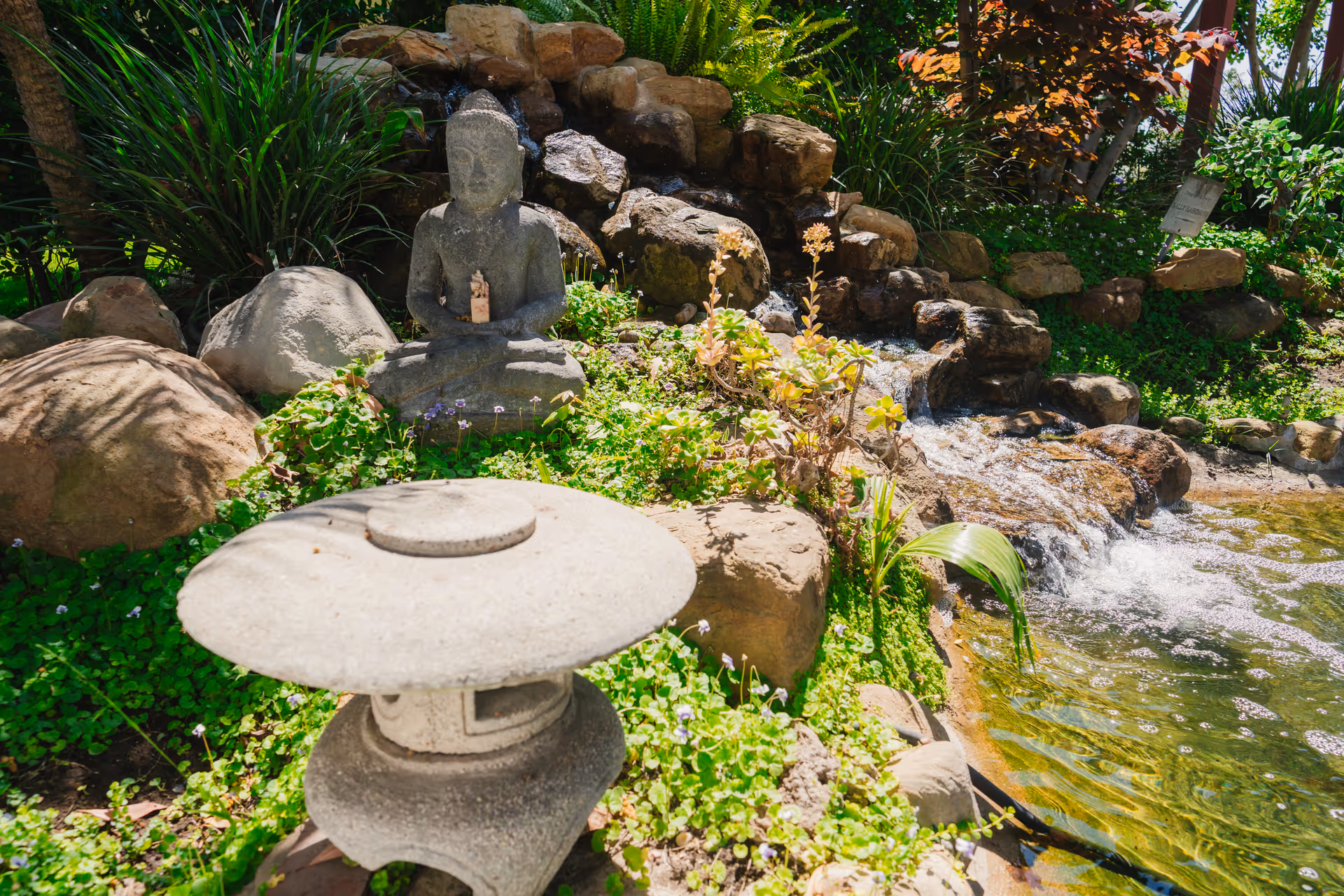 A serene garden scene featuring a stone Buddha statue seated among rocks and greenery next to a small cascading waterfall flowing into a pond. In the foreground, there is a traditional stone lantern surrounded by lush plants and flowers.