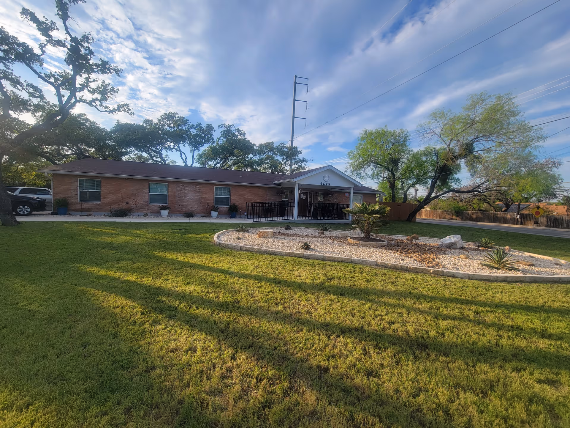 Single-story brick assisted living building with a front porch, landscaped rock bed and a large grassy lawn under a blue sky.