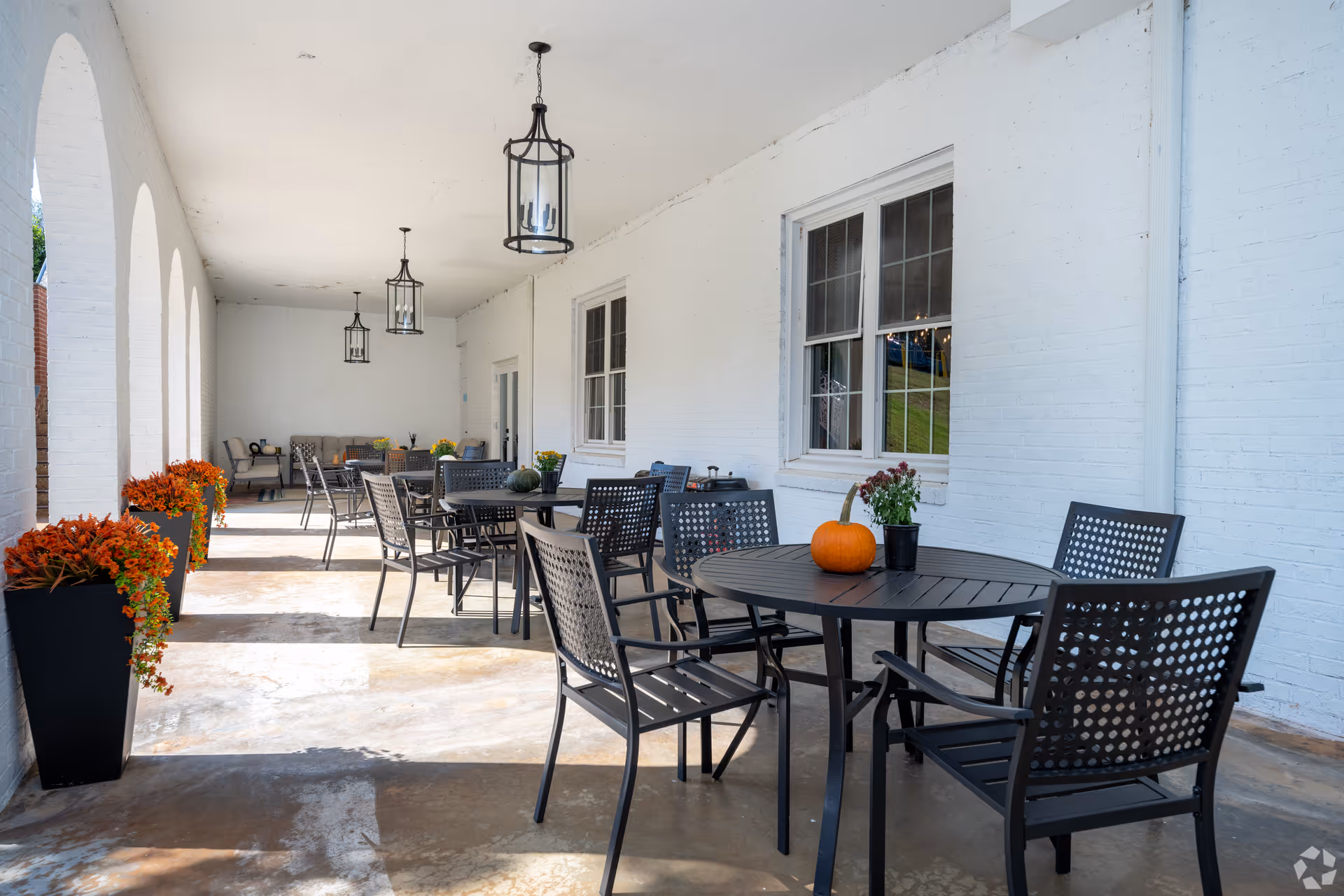 Covered outdoor patio area with black metal tables and chairs, decorated with pumpkins and potted plants. The patio has white brick walls, arched openings on one side, and hanging lantern-style light fixtures on the ceiling.