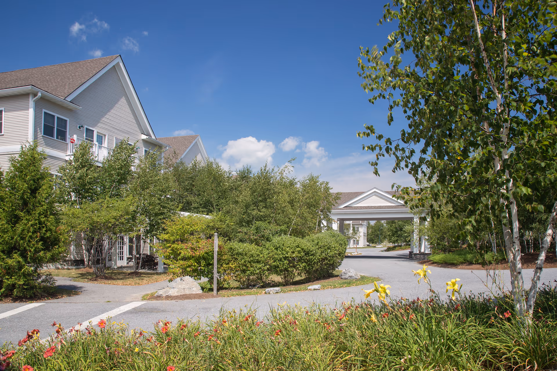 Exterior view of a senior living facility with a driveway, landscaped bushes, trees, and colorful flowers in the foreground. The building has light-colored siding and multiple windows under a clear blue sky with some clouds.