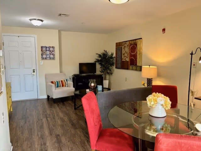 Interior view of a senior living facility room with a glass dining table surrounded by red chairs, a gray sofa, a beige armchair with a colorful pillow, a TV on a stand, a potted plant, wall art, and a lamp on a side table. The floor is wood, and there is a white door in the background.