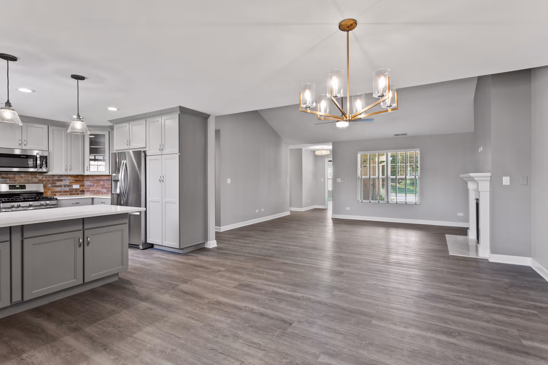 Spacious open-concept interior featuring a modern kitchen with gray cabinets, stainless steel appliances, and a white countertop island. Adjacent to the kitchen is a large living area with wood flooring, a contemporary chandelier, a window with white blinds, and a white fireplace on the right wall.