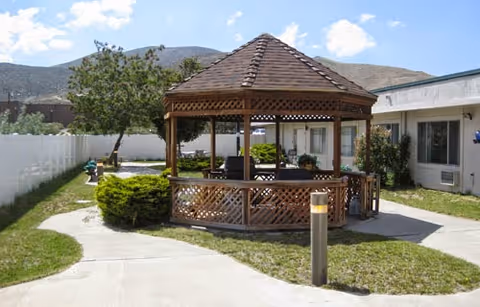 Outdoor area at Mountain View Health and Rehabilitation Center featuring a wooden gazebo with seating, surrounded by a concrete walkway, green grass, bushes, and a white fence with mountains in the background under a partly cloudy sky.